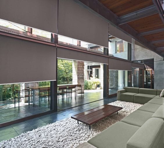 Una sala de estar amplia y luminosa con grandes ventanales que dan a un patio interior arbolado, equipados con persianas enrollables marrones bajadas a media altura. En el interior, se observa un sofá modular gris y una mesa de centro de madera, en un espacio con suelos de piedra y techos de madera.