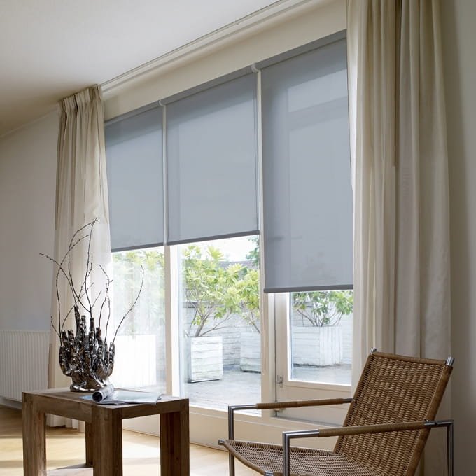 Una ventana grande con tres persianas enrollables grises a media altura, complementadas por cortinas de tela clara colgando a los lados. Se observa una silla de mimbre y madera en el primer plano y vegetación exterior a través de la parte inferior de la ventana.